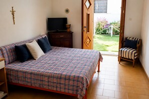 A bedroom with a bed, a television, a wooden cabinet, a chair, and an open door leading to a garden.