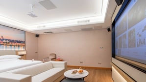 Habitación de hotel con cama, sofá, una mesita con fruta, pantalla de proyector y vistas a la ciudad.