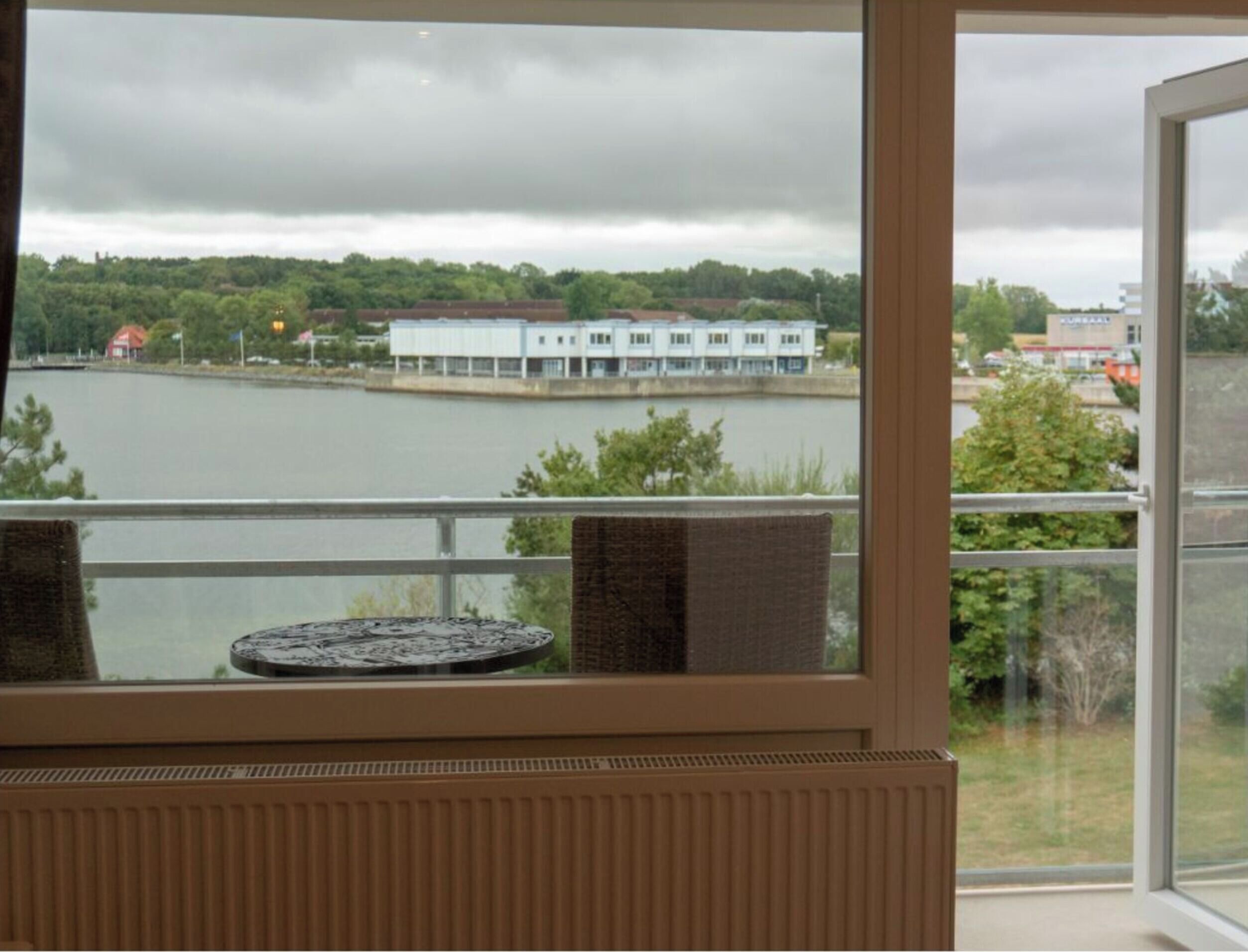 A room with a view of a lake, a building across the water, and a balcony with a table and chairs.