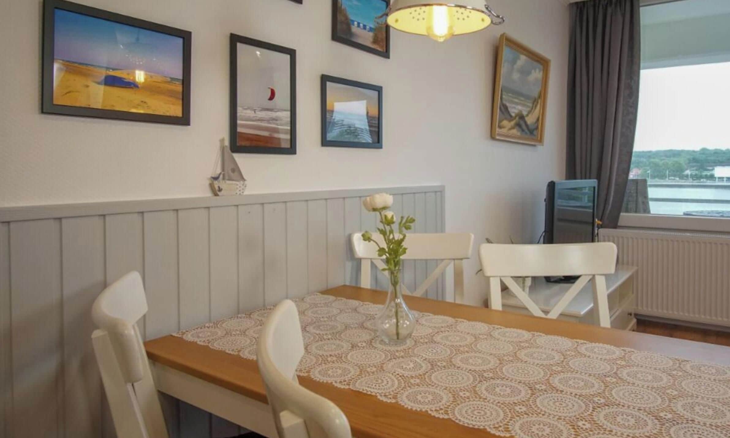 A dining area with a table set for two, a vase with flowers, and a view of a body of water through a window.