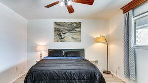 Ein Schlafzimmer mit einem Bett, einem Deckenventilator, einer Wandleuchte, einer Stehlampe und einem Fenster mit Vorhängen.