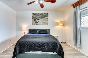 Ein Schlafzimmer mit einem Bett, einem Deckenventilator, einer Wandleuchte, einer Stehlampe und einem Fenster mit Vorhängen.