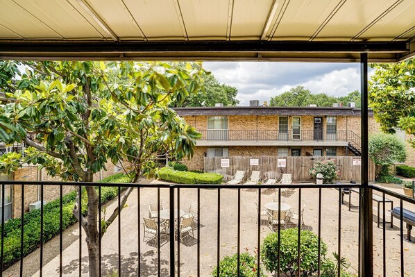 A covered outdoor area with a patio table and chairs, a tree, and a building with a brick facade in the background.