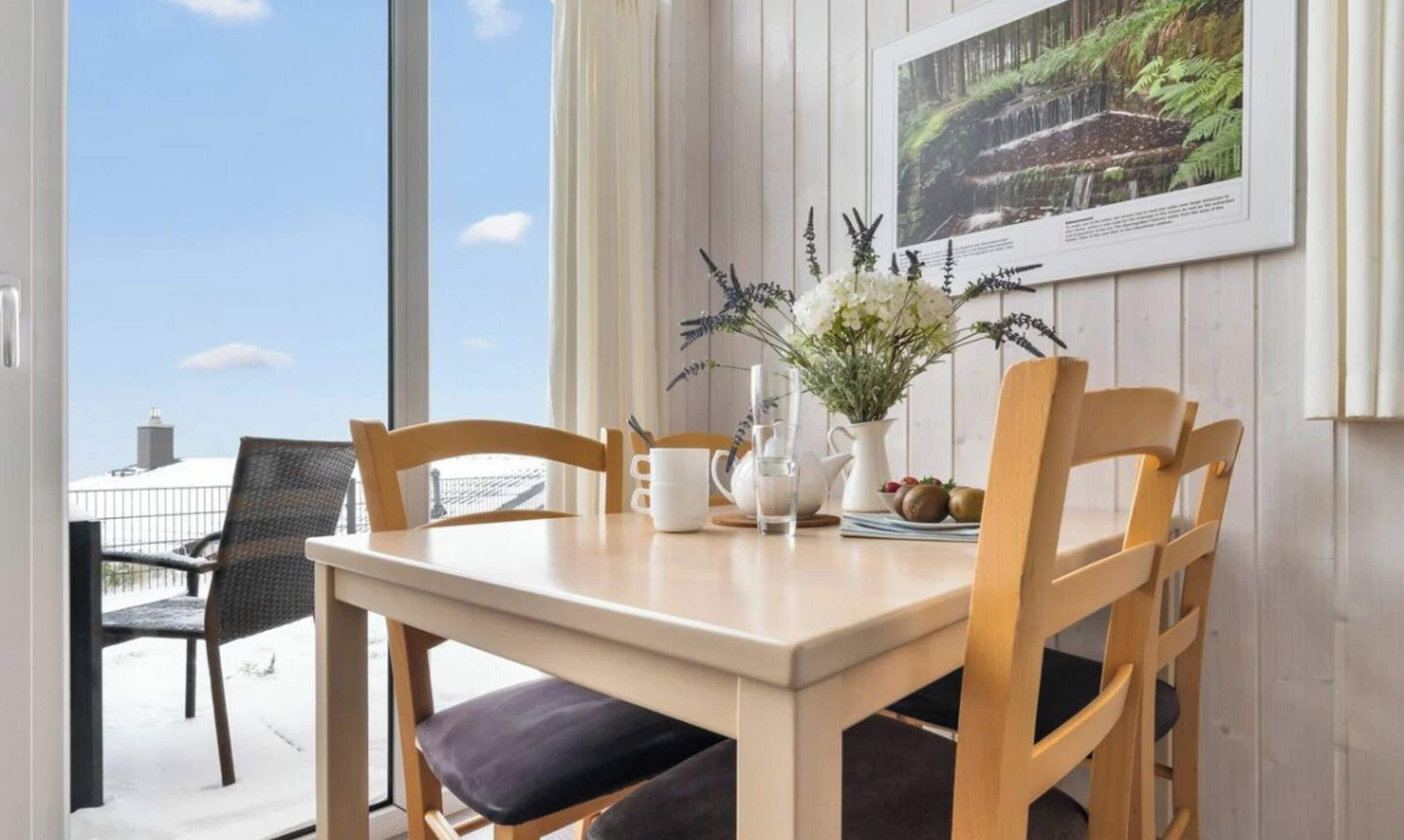 Spisestue med et bord, stole, en vase med blomster, en kande og et billede i ramme på væggen.