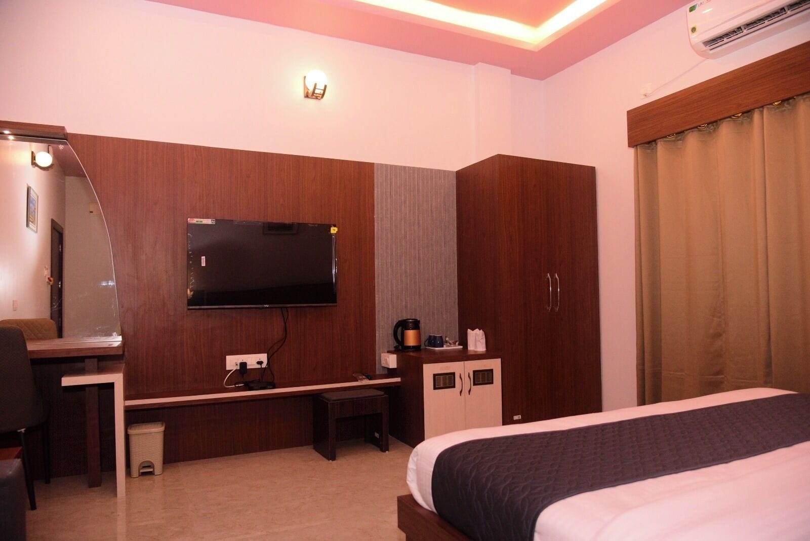 Ein Hotelzimmer mit einem Bett, einem Schreibtisch mit Stuhl, einem an der Wand befestigten Fernseher, einem Kleiderschrank und einer Klimaanlage.
