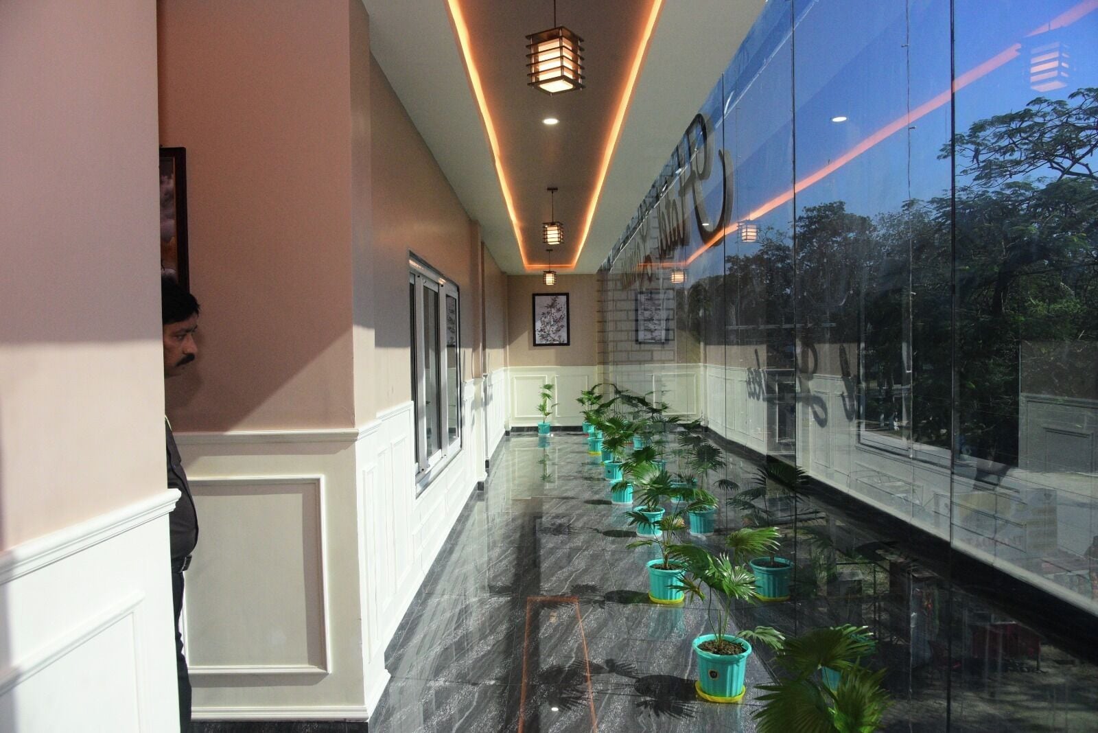 Um corredor com fileira de plantas em vasos, parede de vidro refletindo árvores e teto com iluminação embutida.
