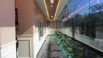Um corredor com fileira de plantas em vasos, parede de vidro refletindo árvores e teto com iluminação embutida.