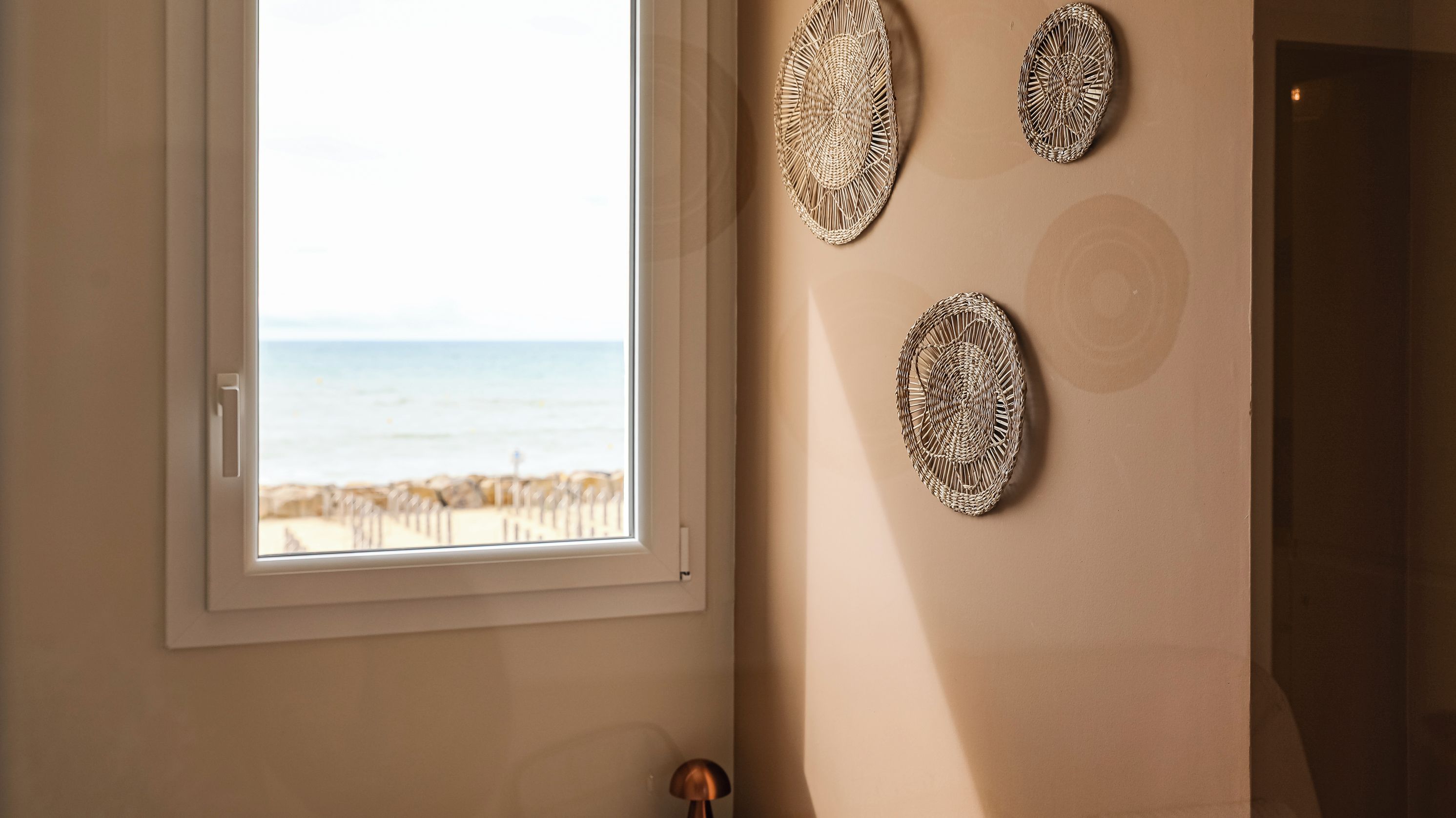 Een kamer met uitzicht op het strand, zichtbaar door een raam en een deur, met drie geweven decoraties aan de muur.