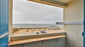 Blick aus einem Fenster auf einen Strand mit einer hölzernen Bank, einem abgestellten Auto und einem Schild.