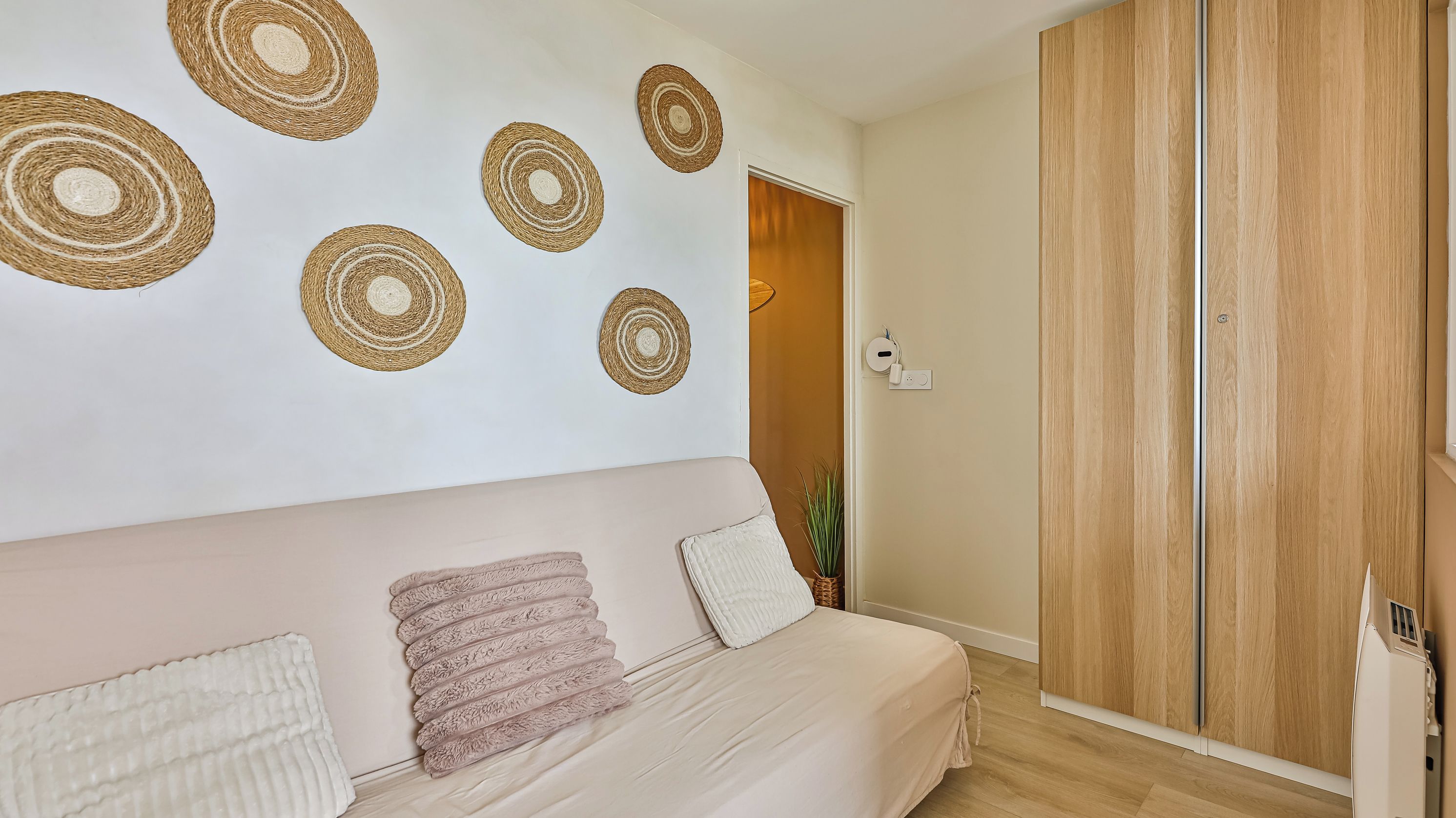 A bedroom with a bed, a wall-mounted circular woven decoration, a wooden wardrobe, and a door leading to another room.