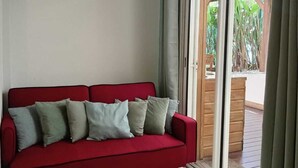 Un divano rosso con numerosi cuscini, un pavimento piastrellato e una porta scorrevole in vetro che dà su una zona esterna con pavimento in legno e vegetazione.