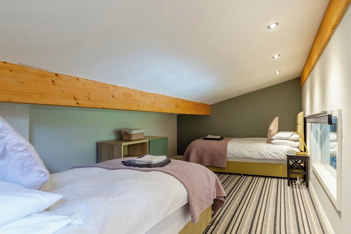 Une chambre d’hôtel avec deux lits, une poutre en bois au plafond, une petite table avec une lampe et une fenêtre donnant sur un beau paysage.
