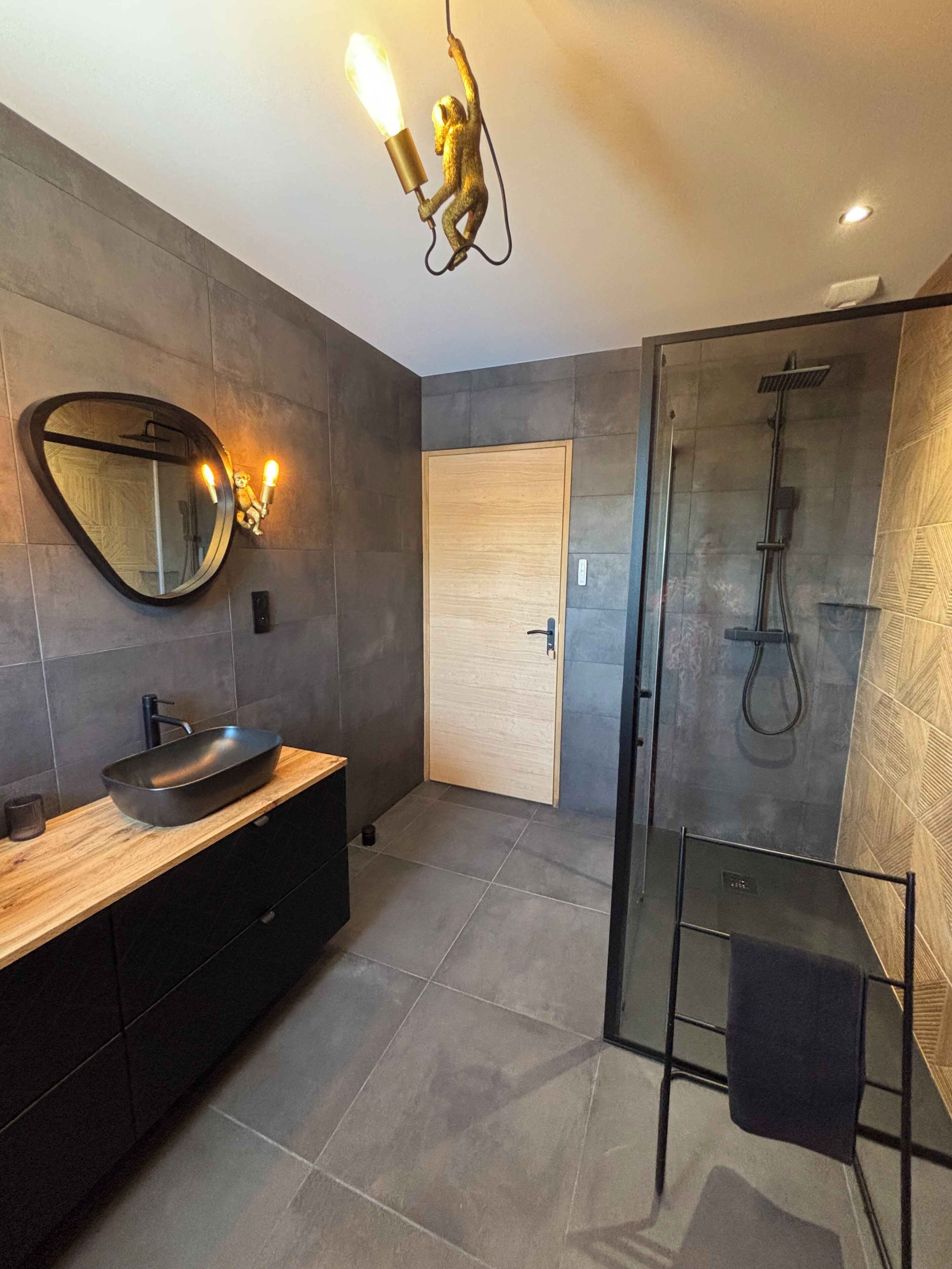 A modern bathroom with a large mirror, a wooden vanity, a black sink, a glass shower enclosure, and a ceiling light fixture.