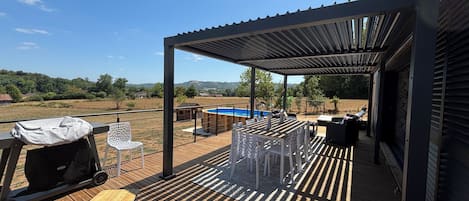 Terraza de madera con mobiliario exterior, una parrilla de barbacoa y vistas a una piscina y colinas.