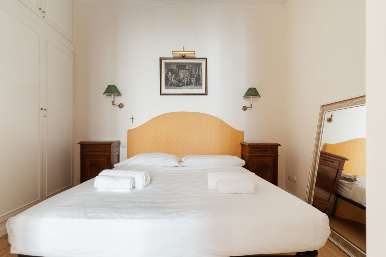 A neatly made bed with white linens and two pillows, a framed picture above the headboard, and two bedside lamps.