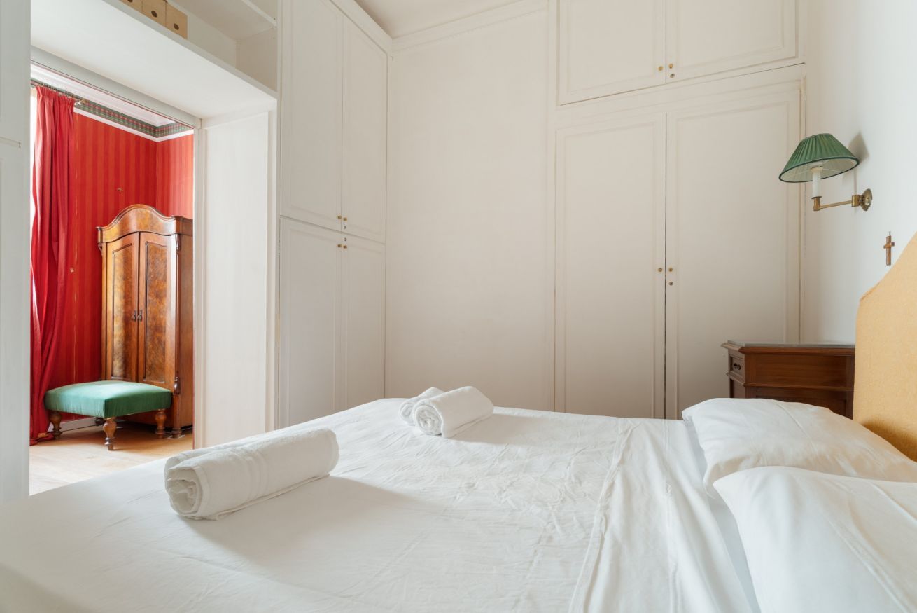 A bedroom with a bed, a wooden wardrobe, a green bench, and a wall-mounted lamp.