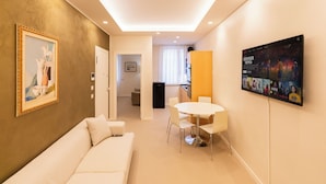 A modern hotel room with a sofa, a dining table with chairs, a kitchen area, and a wall-mounted TV displaying a movie. - Residenza De Nicolis - 50mt Piazza Erbe (Verona)