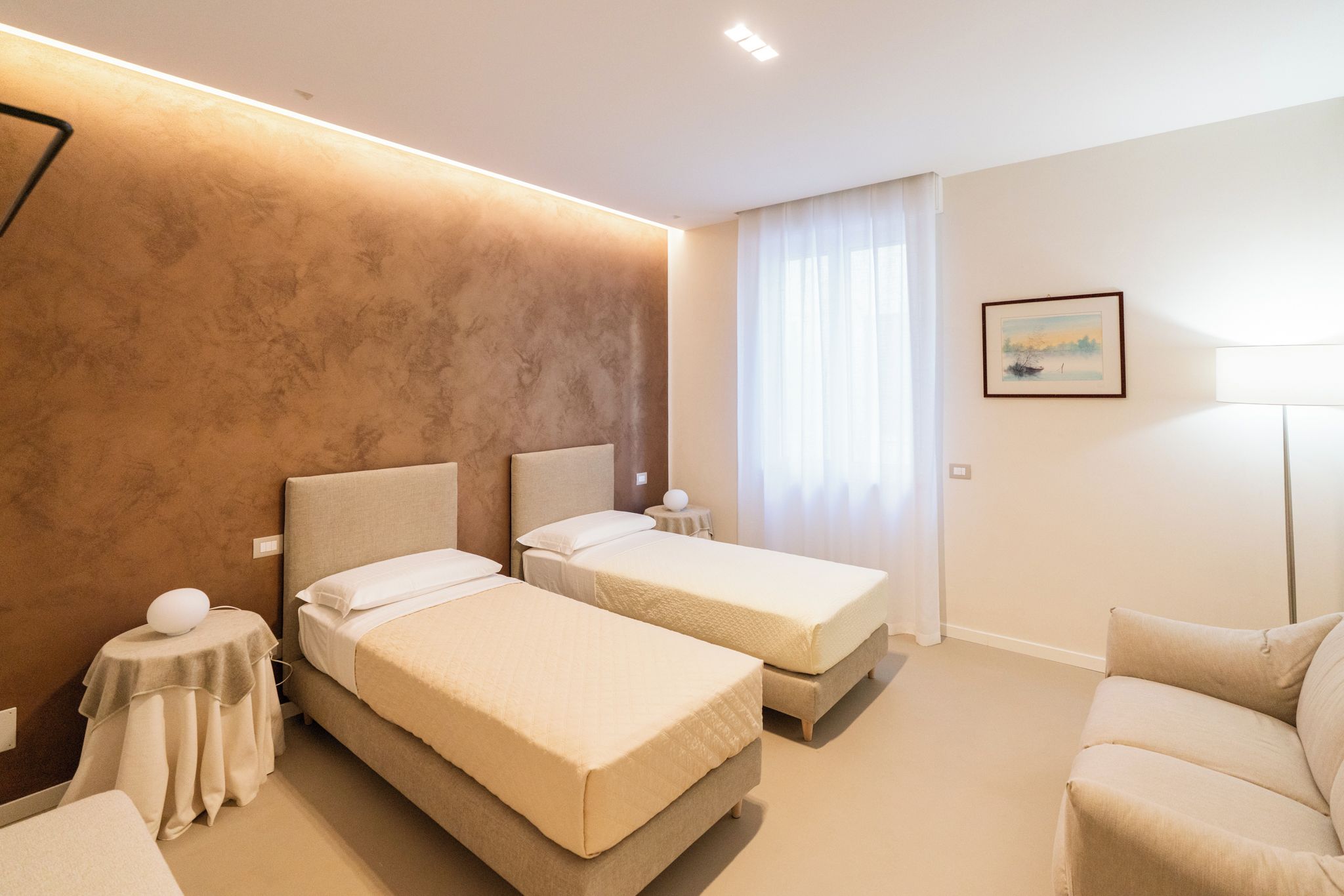 Habitación de hotel con dos camas, un sofá, una lámpara, un cuadro y vistas al exterior.
