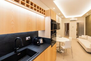 Una cocina moderna con un lavamanos negro, armarios de madera y un horno empotrado. Hay una zona de comedor con una mesa y sillas blancas, y un sofá blanco en la sala de estar.
