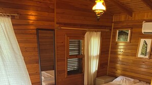 A wooden cabin room with two beds, a mirror, a window with blinds, and a picture on the wall. - Arzum Bungalovs (KUMLUCA)