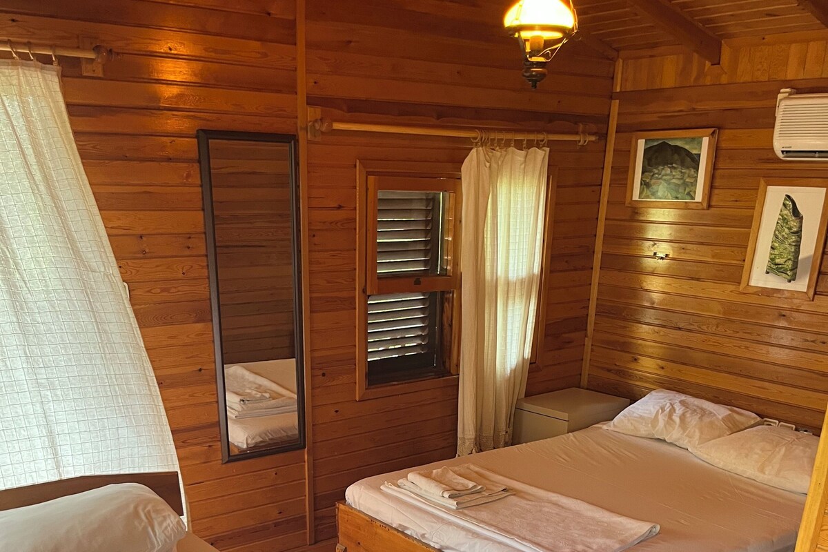 A wooden cabin room with two beds, a mirror, a window with blinds, and a picture on the wall.