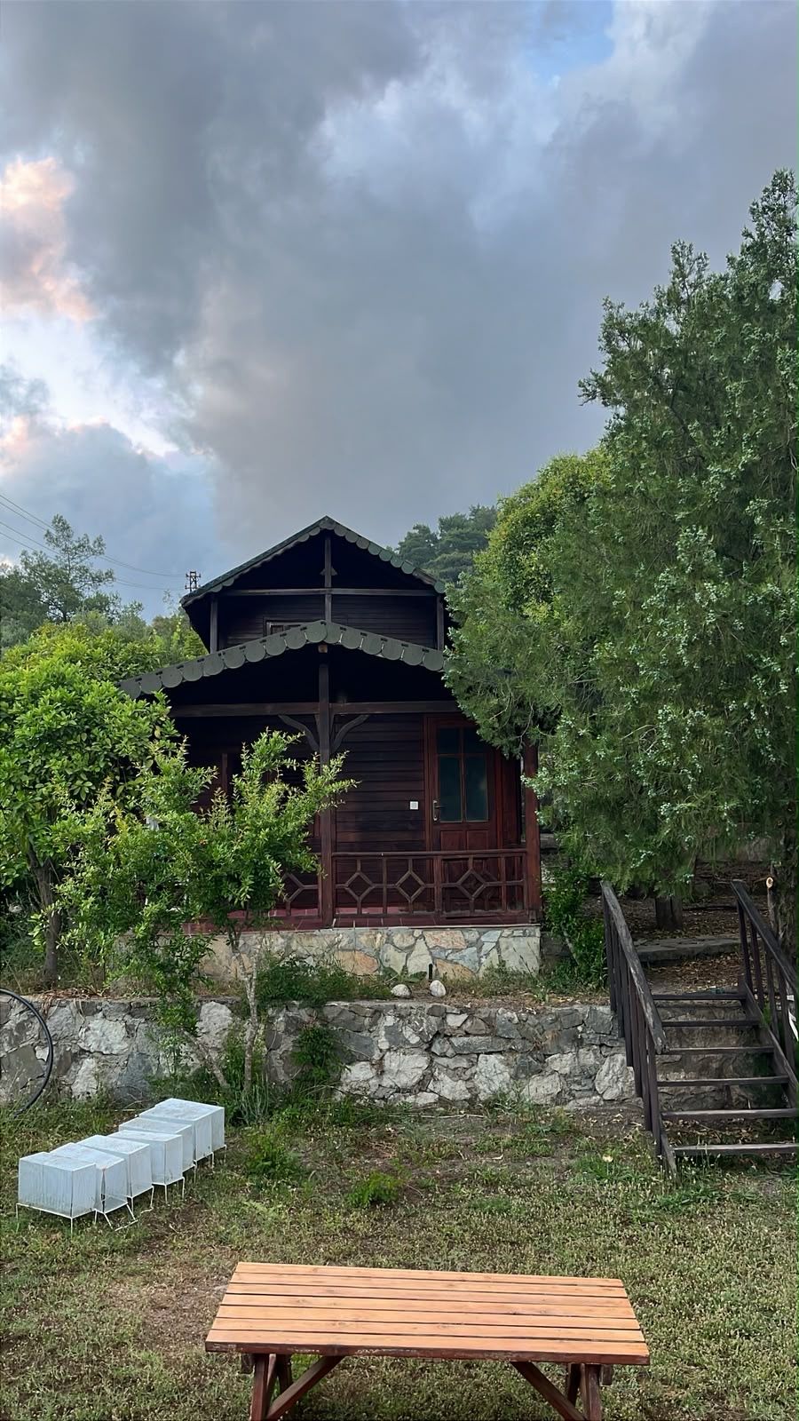Una cabaña de madera con cimientos de piedra, rodeada de vegetación, y un banco de madera en primer plano.