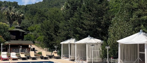 A swimming pool with lounge chairs and canopy tents, surrounded by trees and a hill.