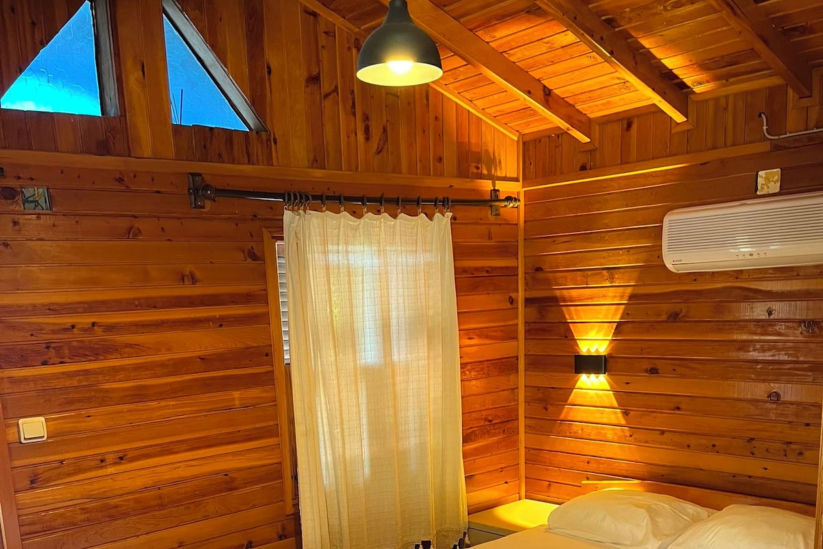 A wooden cabin bedroom with a bed, a hanging light fixture, and a window with a view.