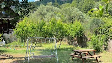 Un parque infantil con tobogán, columpios y una mesa de picnic en un espacio cubierto de césped, con árboles y una montaña al fondo.