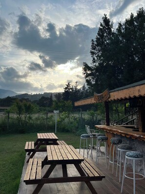 Un área de estar al aire libre con mesas de picnic de madera y taburetes de bar, una barra de madera y vistas a un paisaje montañoso bajo un cielo nublado.