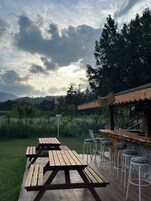 An outdoor seating area with wooden picnic tables and bar stools, a wooden bar, and a view of a hilly landscape under a cloudy sky.