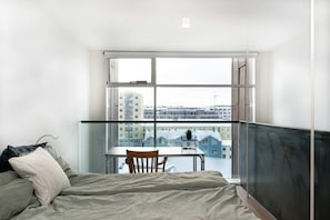 Ein Schlafzimmer mit Bett, Tisch und Stuhl, ein großes Fenster mit Blick auf Gebäude sowie ein Balkon mit Geländer.
