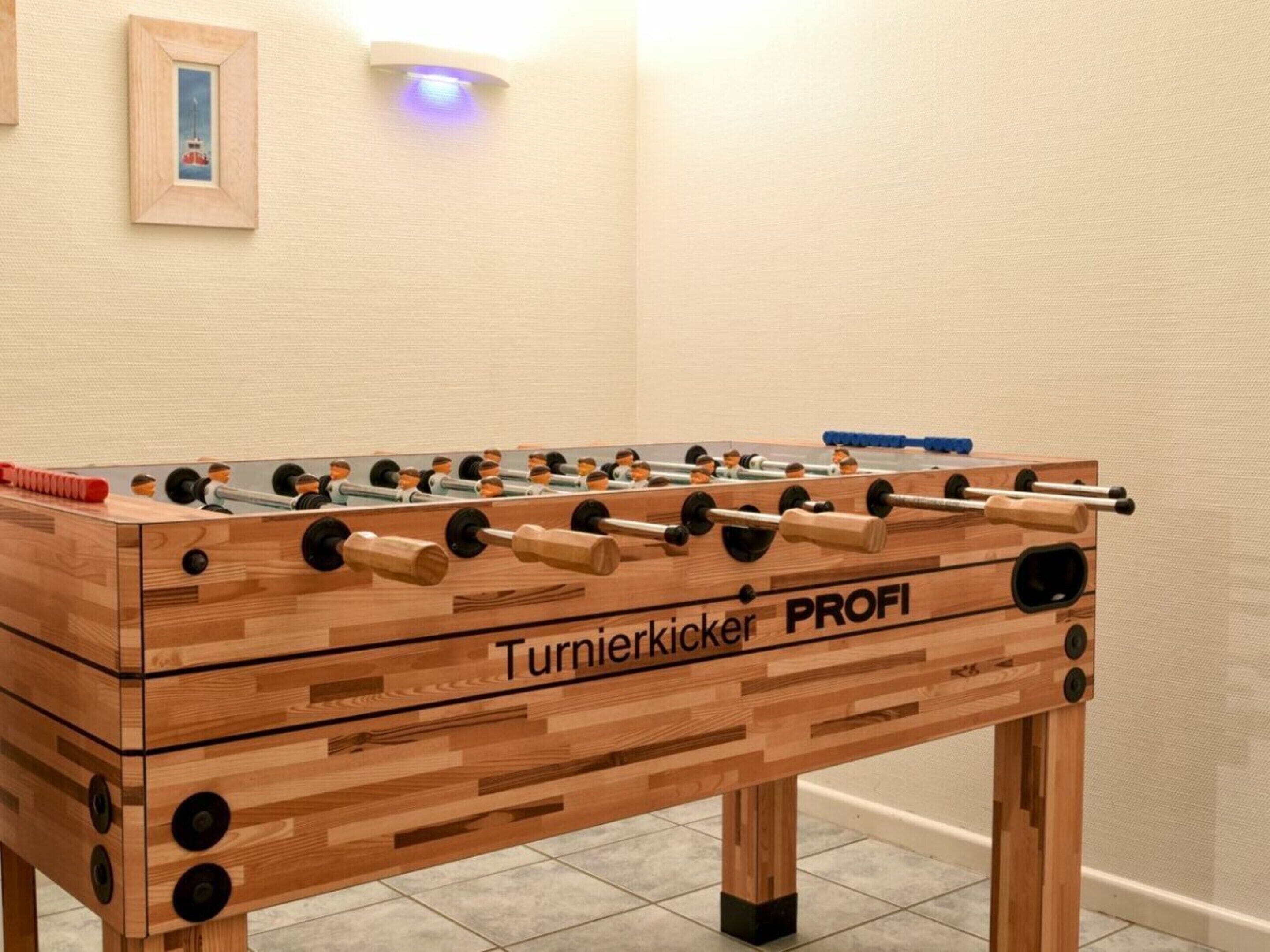 Une table de baby-foot en bois, portant la marque « Turnierkicker PROFI », se trouve dans une pièce dont le sol est recouvert de carreaux et qui est éclairée par un luminaire fixé au mur.