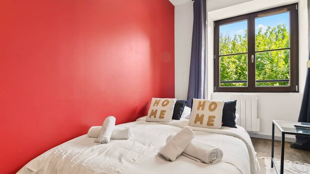 A hotel room with a red wall, a large window with a view of trees, a bed with white bedding and pillows, and a small table. - L'érable - Charming Studio Near Île Barbe (Lyon)