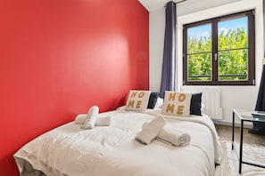 Een hotelkamer met een rode muur, een groot raam met uitzicht op bomen, een bed met witte lakens en kussens, en een klein tafeltje.