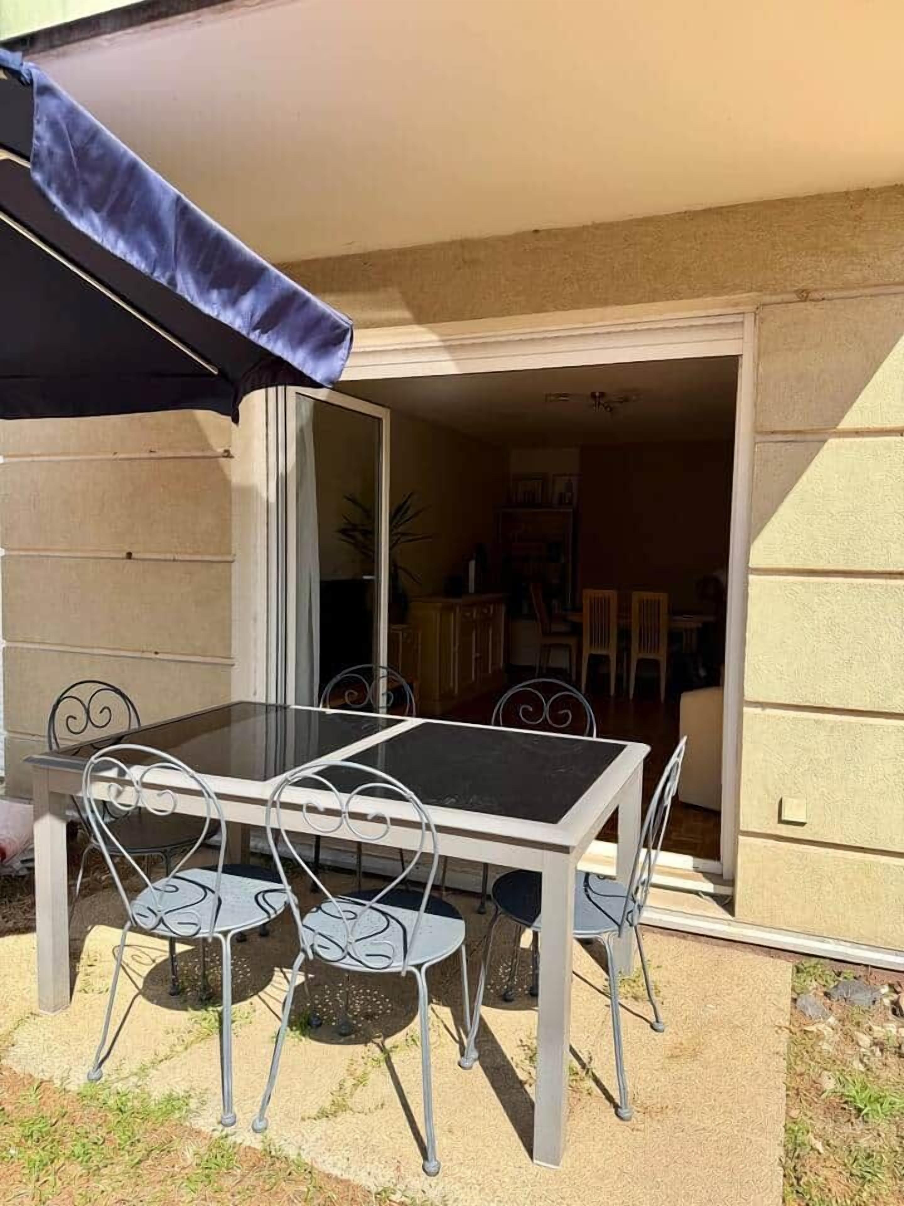 Un patio al aire libre con una mesa y sillas, una puerta corrediza que da a un espacio interior y una vista parcial del exterior de un edificio.