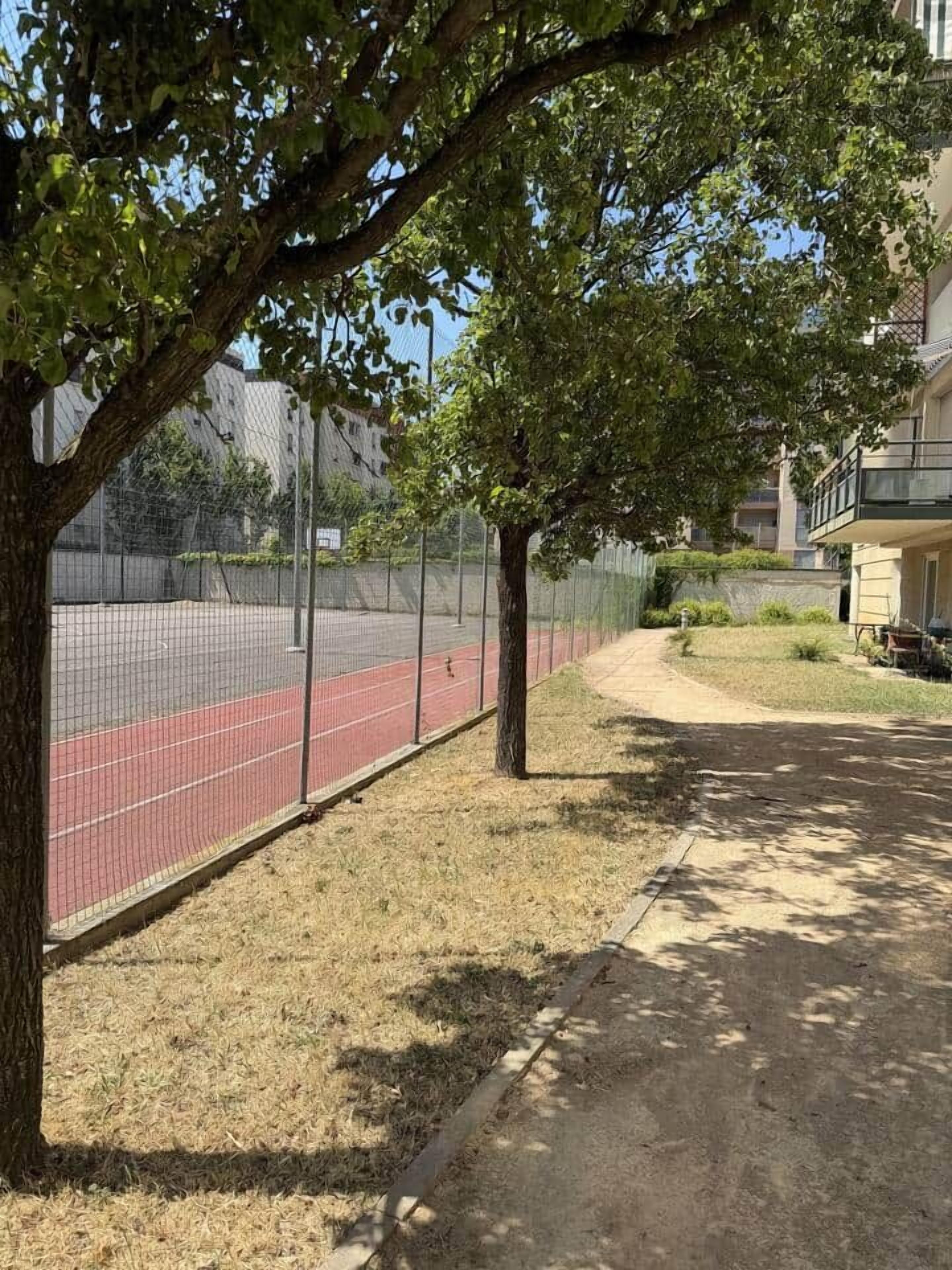 Un sendero conduce a una pista roja, rodeada por una cerca y árboles, con un edificio al fondo.