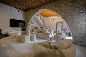A modern living room with a stone archway, a flat-screen TV, and a staircase leading to an upper level. - The Bakery - A Luxury Village Stone House with Pool. Pet Friendly! (Kalavasos)
