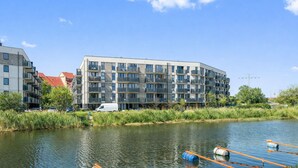 A modern multi-story building with balconies, situated next to a calm river with floating barriers. - Portovo Private Jacuzzi & AC Suite (Gdańsk)