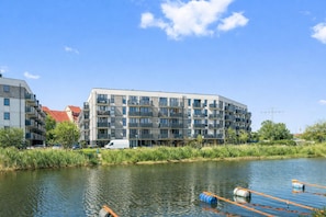 A modern multi-story building with balconies, situated next to a calm river with floating barriers. - Portovo Private Jacuzzi & AC Suite (Gdańsk)