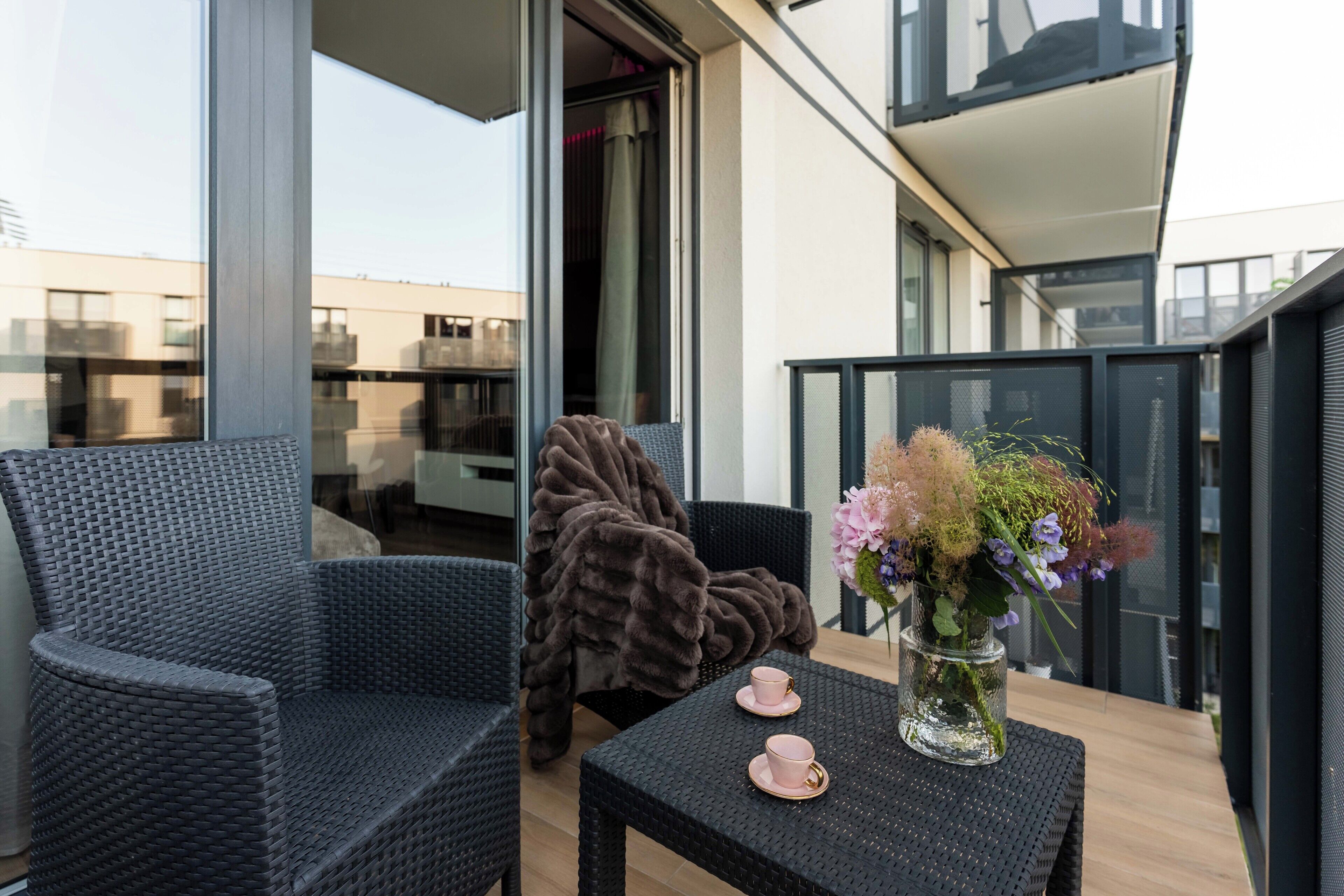 A balcony with wicker furniture, a table with a vase of flowers, and a view of the cityscape.