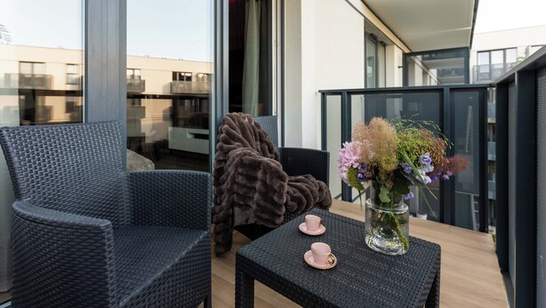 A balcony with wicker furniture, a table with a vase of flowers, and a view of the cityscape. - Portovo Private Jacuzzi & AC Suite (Gdańsk)