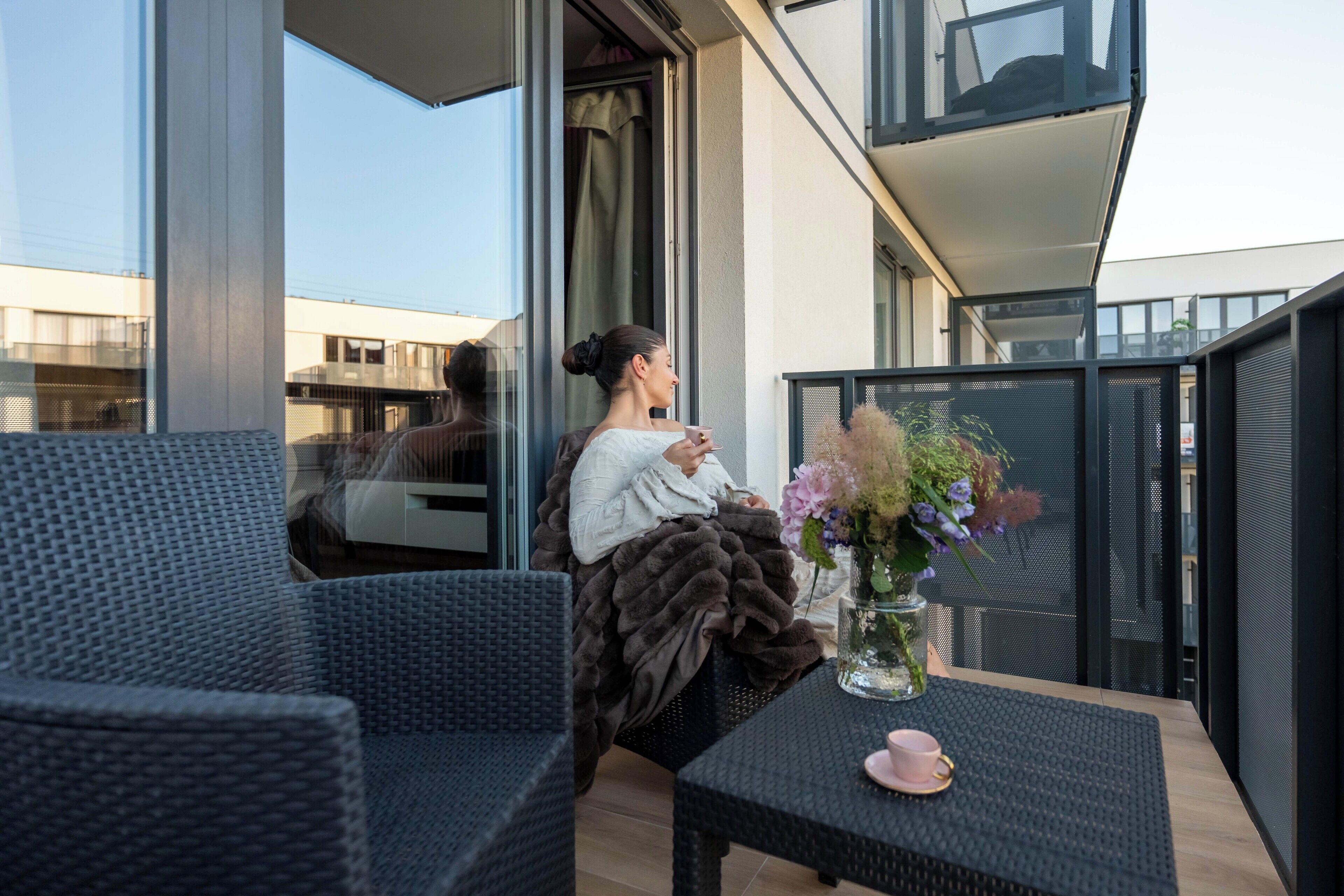 Een balkon met een rieten stoel, een tafel met een bloevas en iemand die zit.