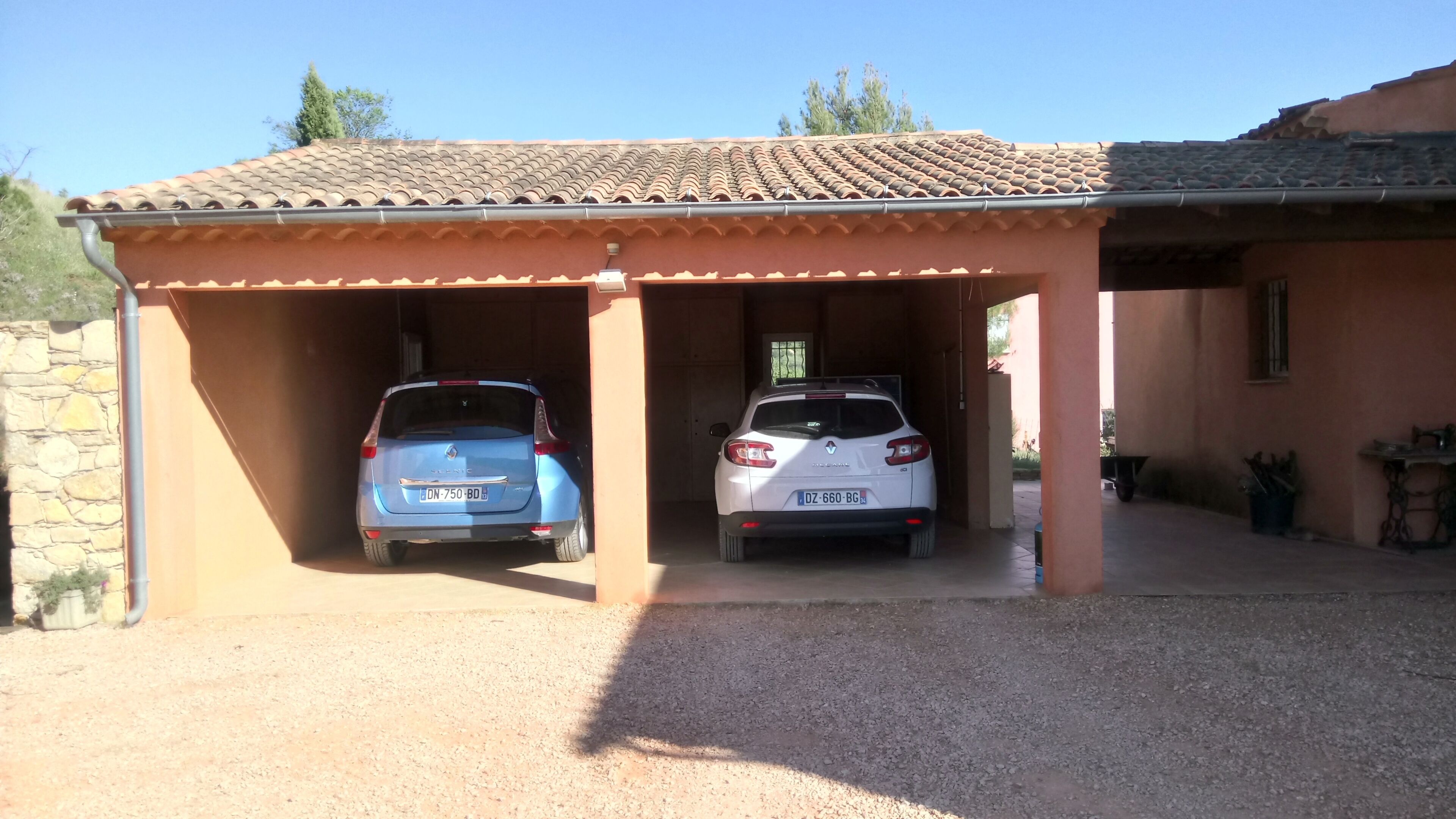 Un espace de stationnement couvert pour voitures, où deux voitures sont garées, avec un mur de pierre à gauche et un bâtiment avec un toit en tuiles à droite.