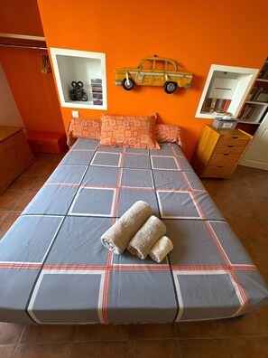 Uma cama com colcha quadricula cinza e laranja, duas toalhas enroladas, uma pequena estante com livros e um carrinho de brinquedo na parede.