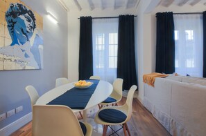 Ein Essbereich mit Tisch, Stühlen und einer Obstschüssel. An der Wand hängt ein Gemälde, und im Hintergrund steht ein Sofa.