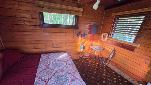 Habitación de cabaña de madera con una cama, una mesita, dos sillas, un ventanal con persianas y un mapa en la pared.