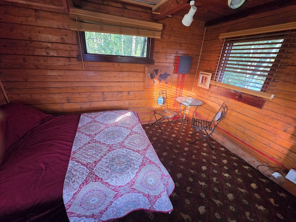 A wooden cabin room with a bed, a small table, two chairs, a window with blinds, and a map on the wall.