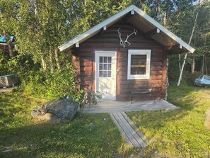 A small log cabin with a wooden porch, a bench, and antlers mounted on the wall.