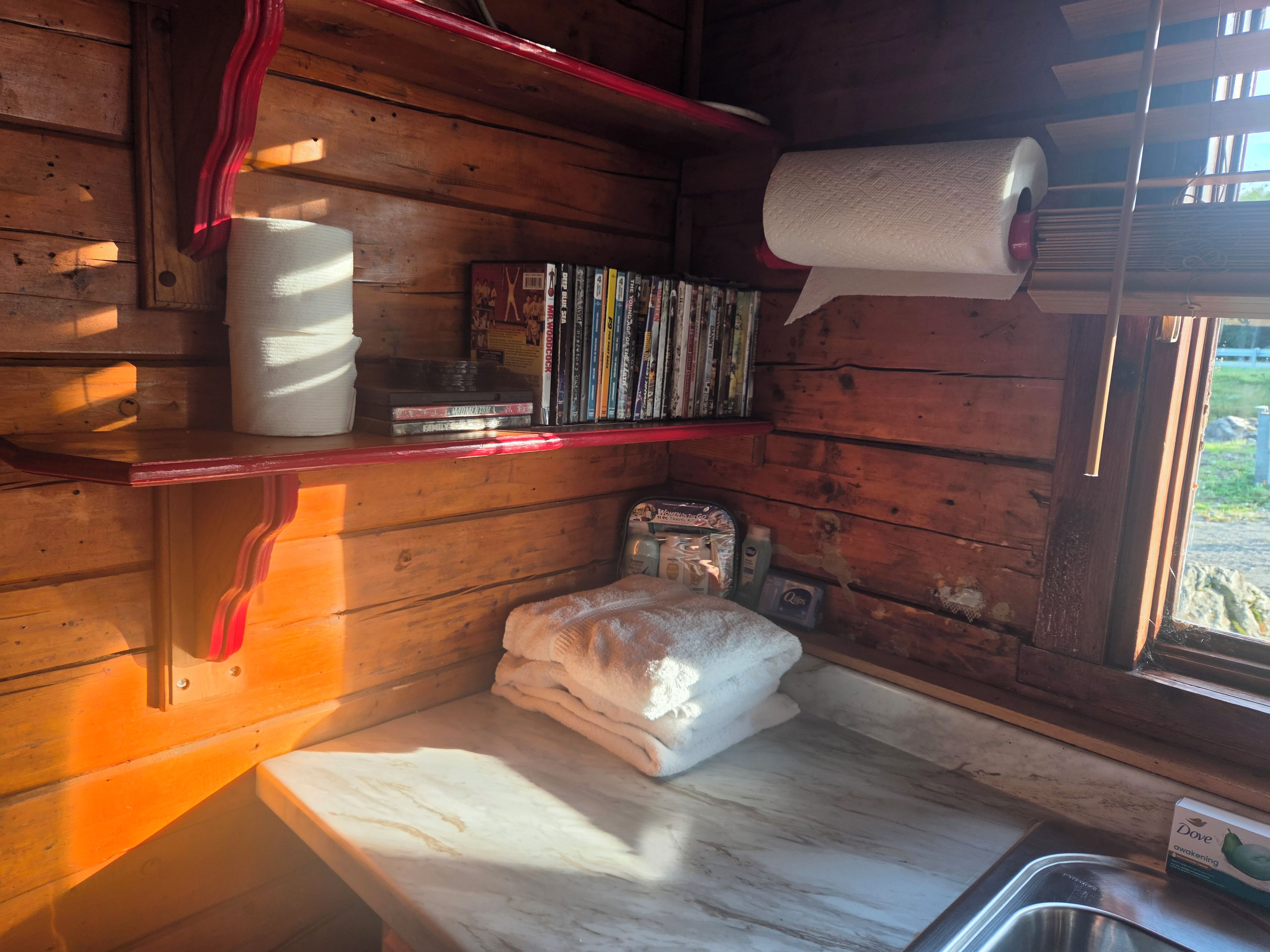 Interior de una cabaña de madera con una repisa que sostiene libros y un rollo de papel higiénico.